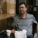 Concentrated Asian male in eyeglasses sitting near window at table with cup of hot drink in outdoor cafe during break