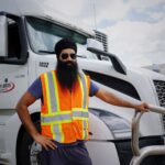 Sikh truck driver with turban and safety vest standing beside a white truck on a sunny day.