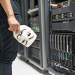 Person holding anonymous mask near servers, hinting at cybersecurity and hacking themes.