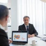 Two businessmen in discussion at an office desk with a laptop.