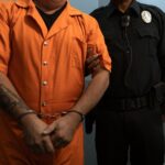 A handcuffed inmate in orange prison uniform being escorted by a police officer.