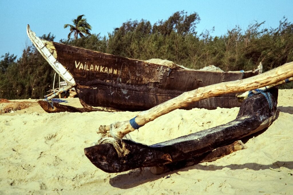 A wooden fishing boat named 'Vailankanni' rests on a sandy beach in India, surrounded by lush trees.