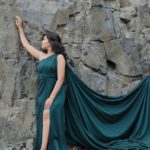 A woman in a flowing green dress poses against a rugged rock wall, exuding elegance and natural beauty.