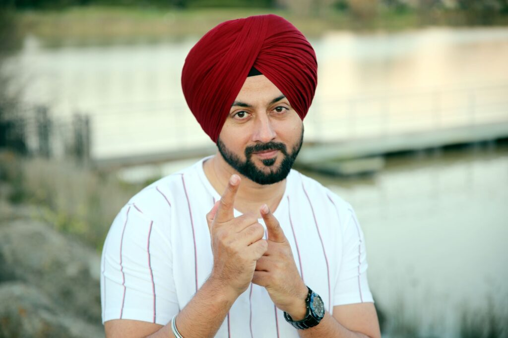 Man wearing red Sikh turban, outdoors near a lake, pointing at camera.