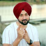 Man wearing red Sikh turban, outdoors near a lake, pointing at camera.