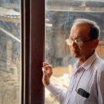 A thoughtful man wearing eyeglasses stands by a glass window, looking outside.