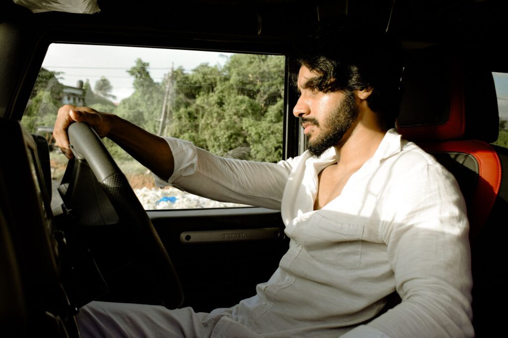 A young man wearing a white shirt drives a car during the day, looking focused.