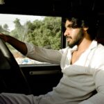 A young man wearing a white shirt drives a car during the day, looking focused.