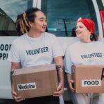 Two smiling volunteers carrying boxes labeled food and medicine by a supply van.