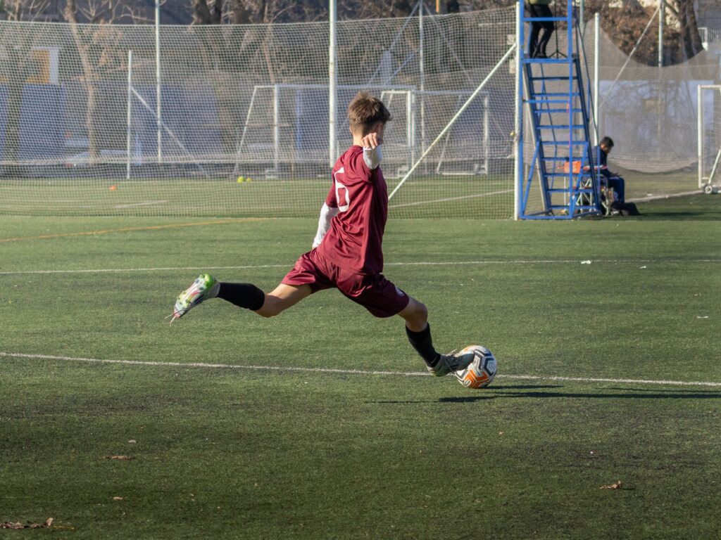 Teenager in action kicking a soccer ball during an outdoor match on a sunny day.