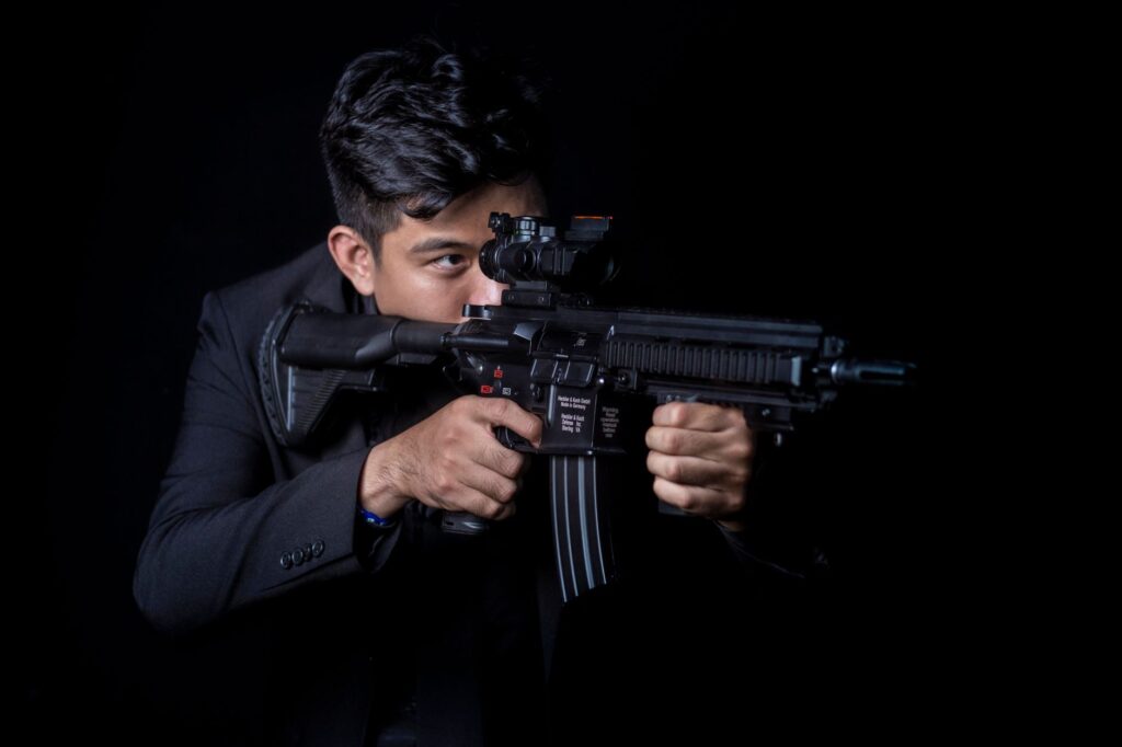 Portrait of a man aiming a rifle with a focused expression against a black background.
