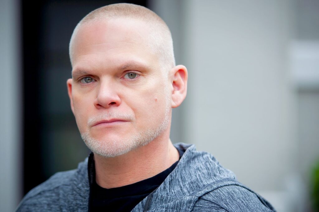 A serious-looking bald man with stubble, photographed outdoors in natural daylight.