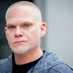 A serious-looking bald man with stubble, photographed outdoors in natural daylight.