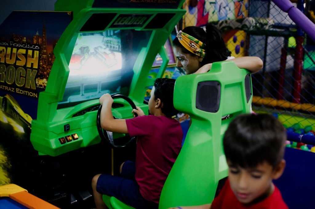 Unrecognizable child having fun with bright car racing simulator while turning steering wheel near serious ethnic woman and funny brother in shopping center near playground