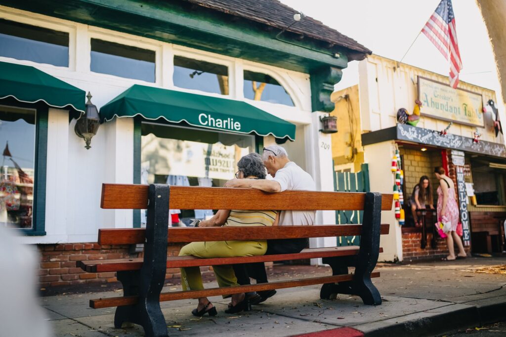 A senior couple sits together on a bench outside a shop, embracing under an American flag.