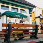 A senior couple sits together on a bench outside a shop, embracing under an American flag.