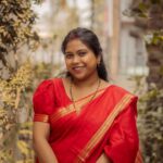 Portrait of a woman wearing a red sari with a soft smile, surrounded by greenery in an outdoor setting.