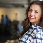 Portrait of a smiling woman in a plaid shirt leaning against a wall indoors.