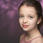 Portrait of calm young girl with bright eyes and hairstyle in pink top looking at camera against purple background