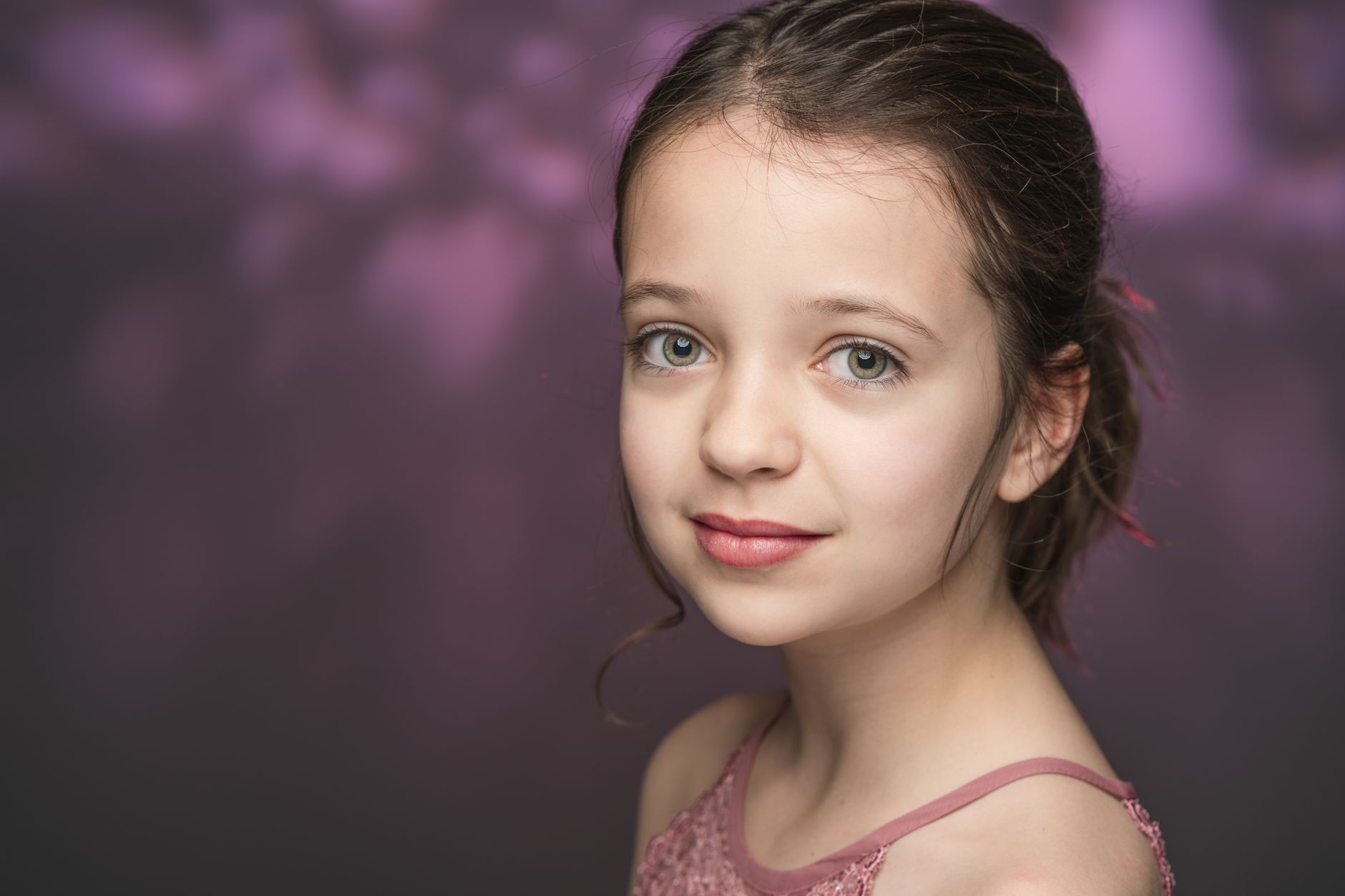 Portrait of calm young girl with bright eyes and hairstyle in pink top looking at camera against purple background