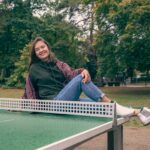 A cheerful woman poses on a ping pong table outdoors in a park setting, surrounded by lush greenery.