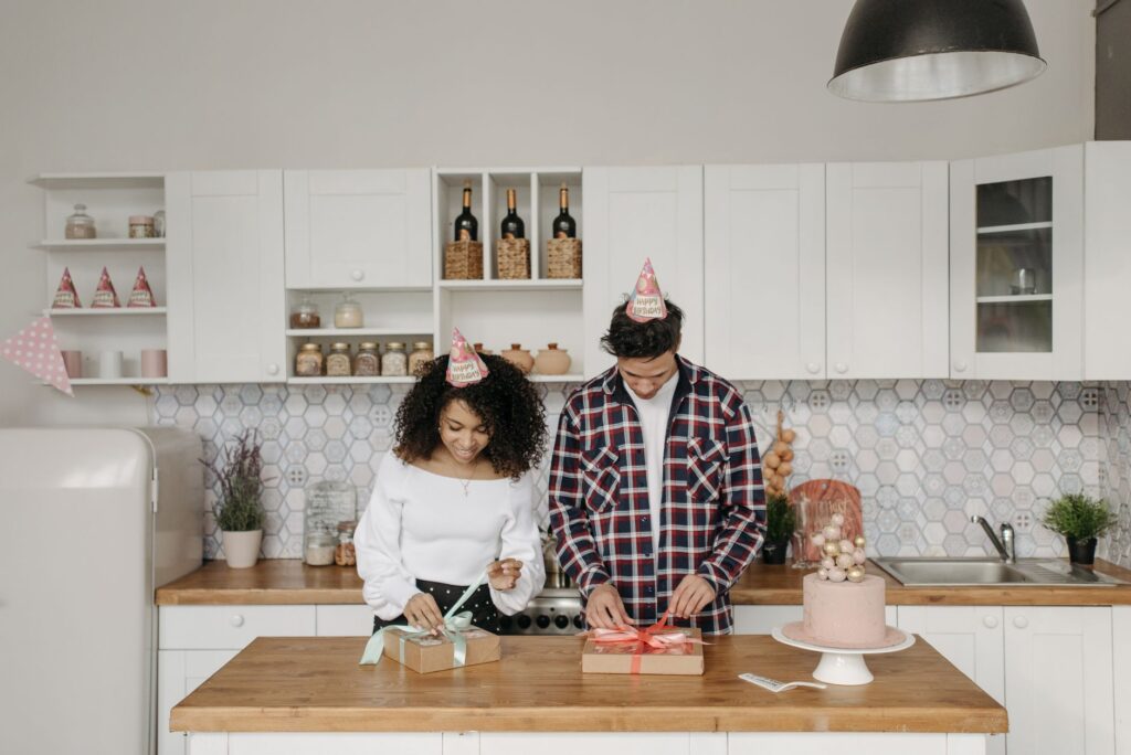 A couple in party hats wrap presents on a kitchen island during a celebration.