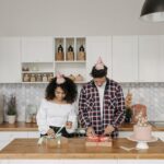 A couple in party hats wrap presents on a kitchen island during a celebration.
