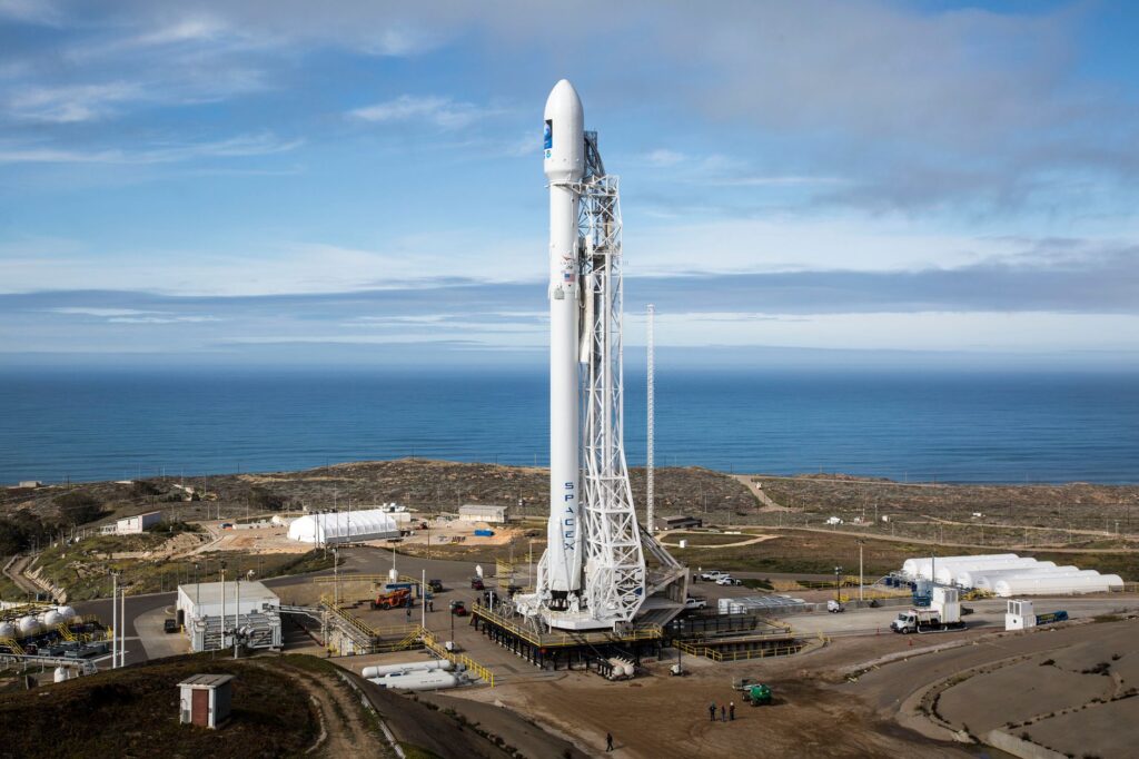 A SpaceX rocket stands on a launch pad overlooking the ocean under a clear sky.