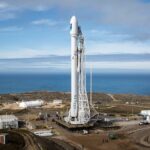 A SpaceX rocket stands on a launch pad overlooking the ocean under a clear sky.