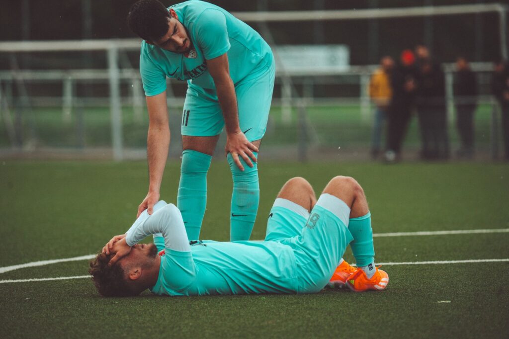 A soccer player lying on the grass field being consoled by a teammate in sportswear.
