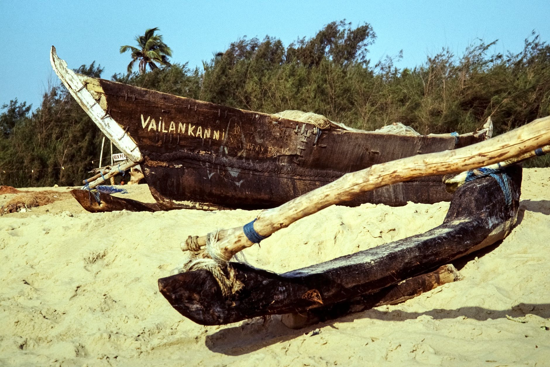 A wooden fishing boat named 'Vailankanni' rests on a sandy beach in India, surrounded by lush trees.