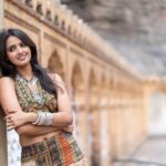 A woman in vibrant ethnic attire with silver jewelry poses by a historic wall.