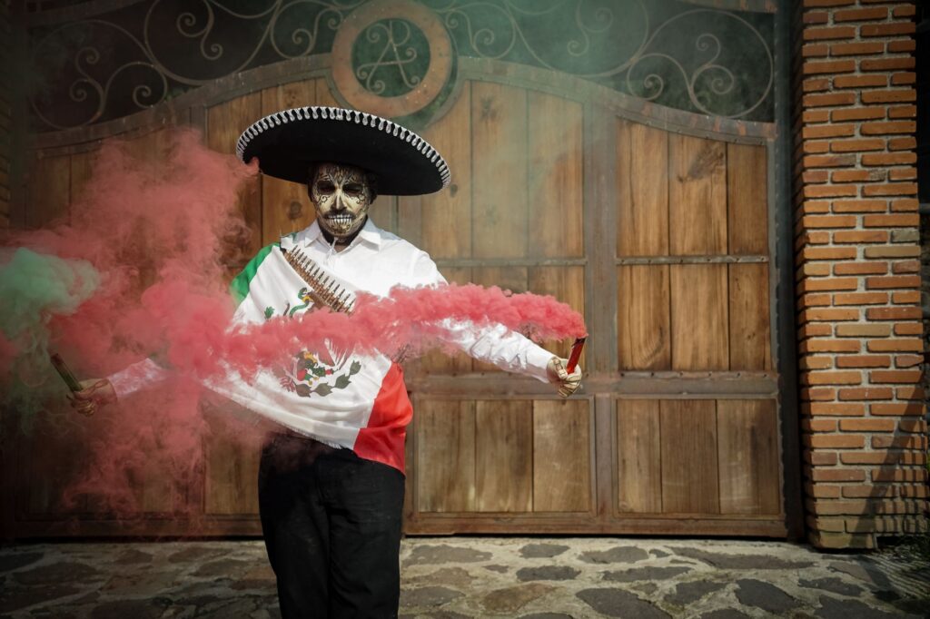 Man in Mexican costume with Dia de los Muertos makeup celebrating with colorful smoke outdoors.