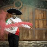 Man in Mexican costume with Dia de los Muertos makeup celebrating with colorful smoke outdoors.