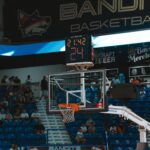 Close-up of a basketball hoop inside a stadium with a scoreboard and audience in the background.