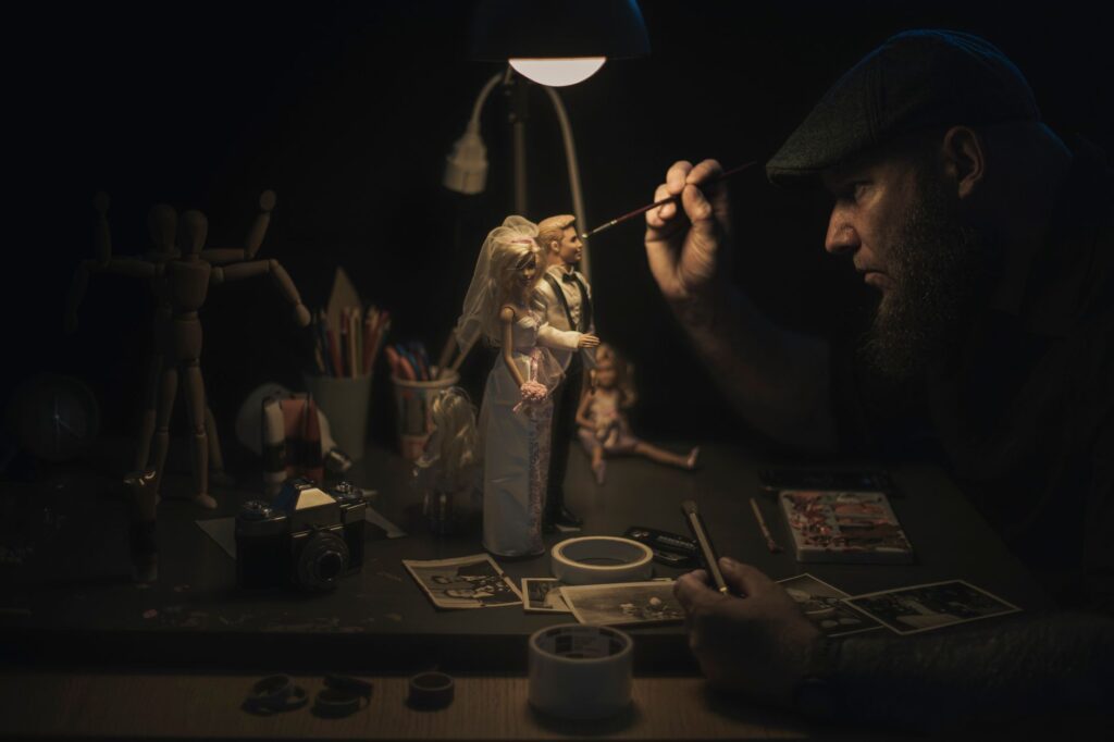 A craftsman meticulously painting bride and groom figurines under a lamp, focused on details.