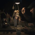 A craftsman meticulously painting bride and groom figurines under a lamp, focused on details.