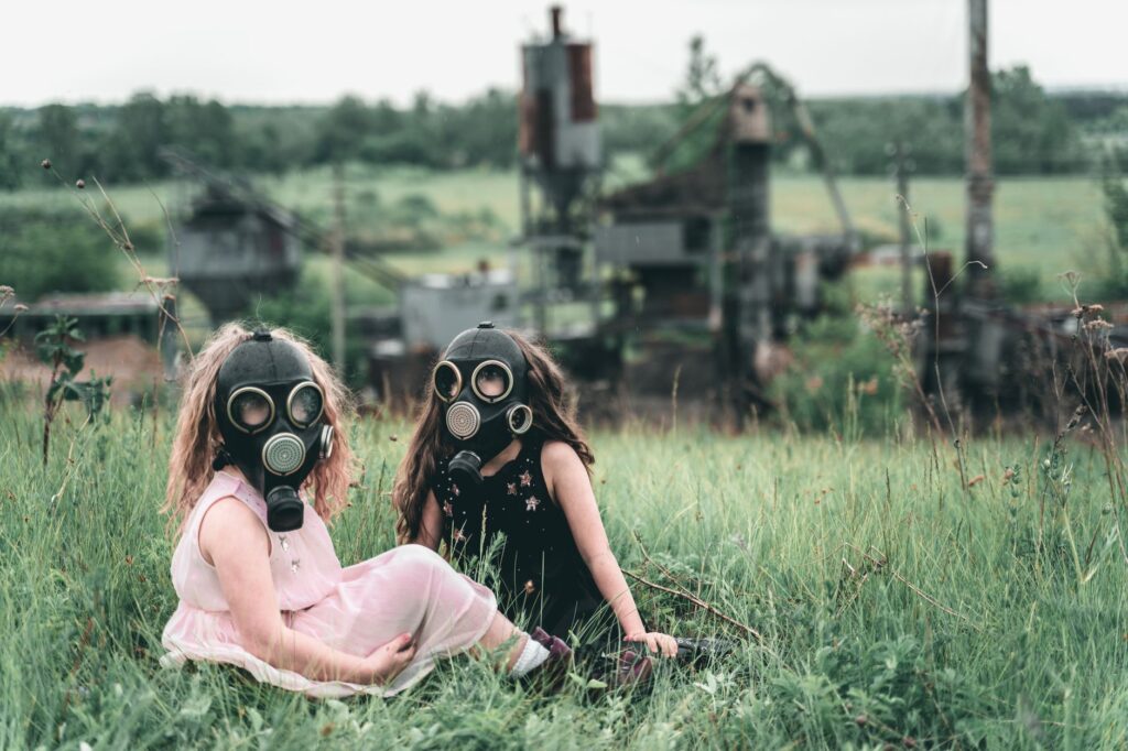 Two children wearing gas masks seated in a grassy field near an abandoned industrial site.