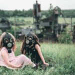 Two children wearing gas masks seated in a grassy field near an abandoned industrial site.