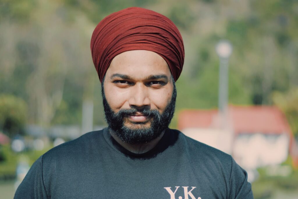 A cheerful man wearing a red turban and black t-shirt enjoys a sunny day outdoors.