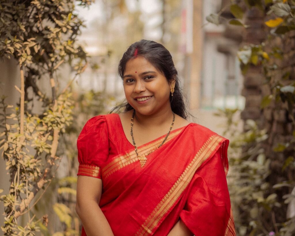 Portrait of a woman wearing a red sari with a soft smile, surrounded by greenery in an outdoor setting.