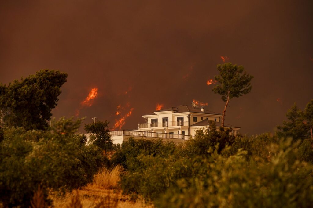 A residential building surrounded by blazing forest fire under a dramatic sky.