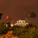 A residential building surrounded by blazing forest fire under a dramatic sky.