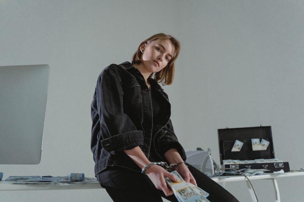 Young woman in black jacket handcuffed with cash, conceptual indoors setting.