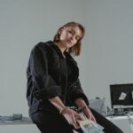 Young woman in black jacket handcuffed with cash, conceptual indoors setting.